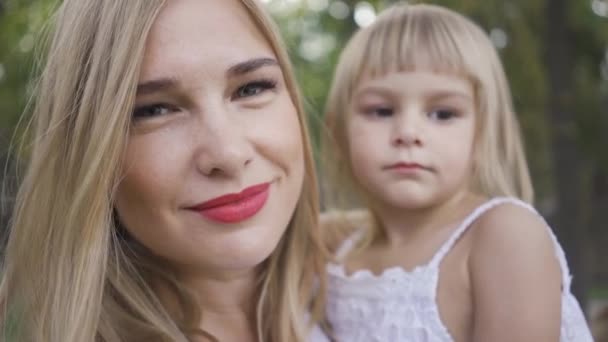 Ritratto di giovane donna bionda che tiene in braccio la sua graziosa figlia vestita di bianco guardando la macchina fotografica. Tempo libero all'aperto. Una famiglia felice. Un genitore solo. Maternità, genitorialità, infanzia felice — Video Stock