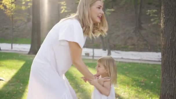 年轻的金发女人旋转着抱着她可爱的女儿在白色礼服的肖像。户外休闲。幸福的家庭一个父级。母性、父母、快乐的童年 — 图库视频影像