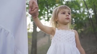 Küçük sarışın kız parkta yürüyen annelerin elini tutarak ve etrafa bakıyor. Mutlu bir aile. Bir ebeveyn. Annelik, ebeveynlik, çocukluk. Alt görünüm