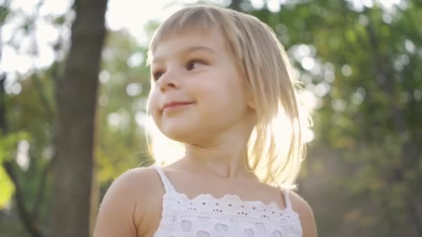 Primer plano de una niña linda mirando a la cámara y apuntando hacia otro lado. Retrato de un niño adorable en el parque bajo los rayos del sol . — Vídeo de stock