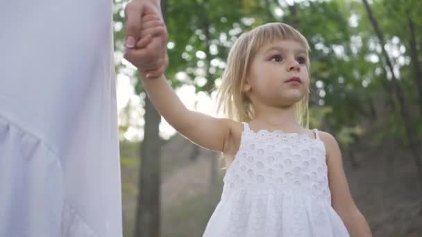 Mała blond dziewczyna chodzenie w parku trzymając matek rękę i rozglądając się. Szczęśliwa rodzina. Jeden rodzic. Macierzyństwo, rodzicielstwo, dzieciństwo. Widok z dołu — Wideo stockowe