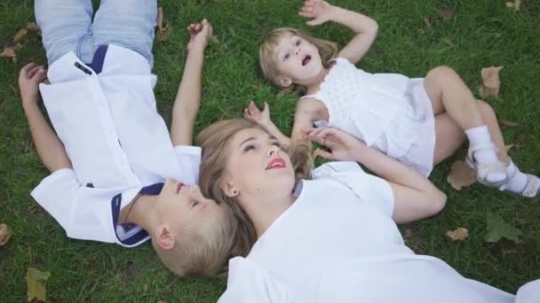 Die hübsche blonde Frau und ihre beiden Kinder liegen auf dem grünen Rasen im Park. Junge, Mädchen und attraktive Frau verbringen Zeit im Freien. Porträt einer glücklichen Familie mit zwei Kindern. Ansicht von oben — Stockvideo