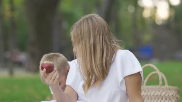 Mulher caucasiana atraente sentado na grama com o menino, segurando maçã na mão. A mãe e o filho passam tempo juntos ao ar livre. Um dos pais. Maternidade, paternidade, infância — Vídeo de Stock