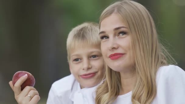 Attraktiv blond kvinna sitter med liten pojke, hålla äpple i handen och pratar. Mor och son spenderar tid tillsammans utomhus. Moderskap, föräldraskap, barndom. — Stockvideo