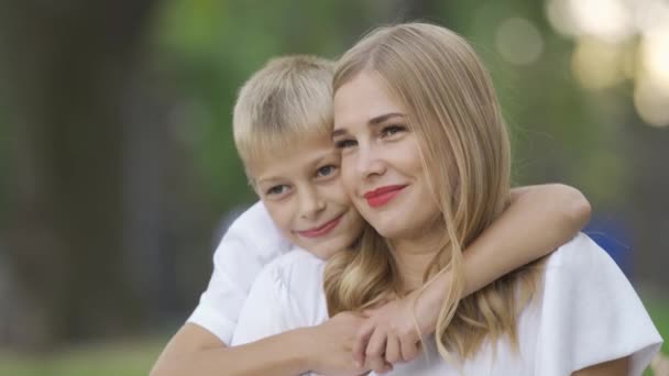 Porträtt av ung kaukasisk kvinna och liten pojke som sitter i parken eller skogen. Sonen kramar sin mor. Lycklig familj umgås tillsammans. Solig dag utomhus. — Stockvideo