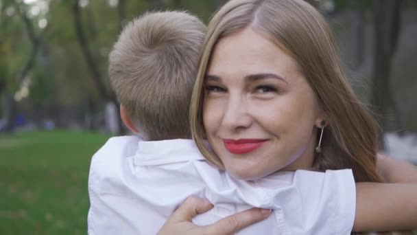 Πορτραίτο μιας ελκυστικής λευκής γυναίκας που κοιτάζει την κάμερα, αγκαλιάζει το μικρό ξανθό αγόρι και χαμογελάει. Η μητέρα περνάει χρόνο με το γιο της στο πάρκο. Αργή κίνηση. — Αρχείο Βίντεο