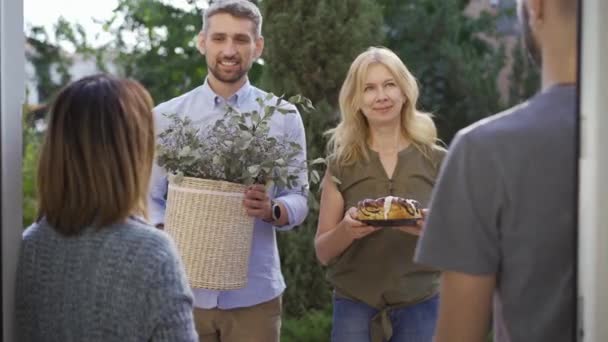 Familia caucásica dando regalos a sus nuevos vecinos. Amistosa pareja saludando a los recién llegados con un pastel y un cubo de flores . — Vídeos de Stock