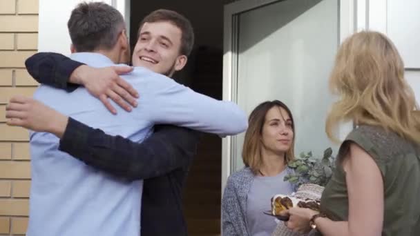 Dwóch ludzi pozdrowienie siebie, podczas gdy ich żony stojących obok nich i mówić. Młoda para kaukaski spotkanie przyjaciół w drzwiach. — Wideo stockowe