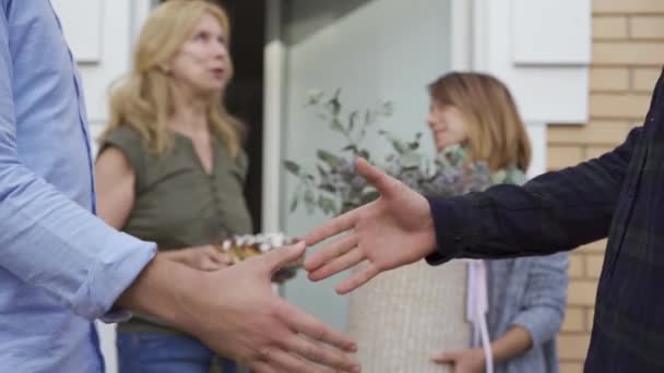 Close up de dois homens apertando as mãos em primeiro plano, enquanto suas esposas conversando no fundo. Conheça novos vizinhos. Conceito de amizade, bom relacionamento — Vídeo de Stock