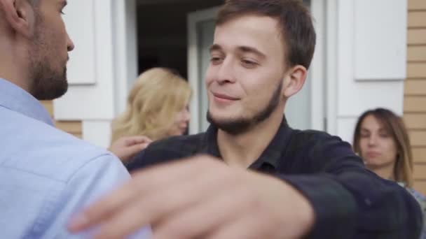 Zwei kaukasische Männer im Vordergrund, die einander grüßen und lächeln. zwei attraktive Frauen im Hintergrund reden und halten eine Torte und einen Blumenkübel in der Hand. — Stockvideo