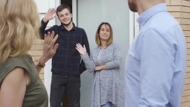 Glückliche Familie, die vor der Tür steht und winkt, während ihre Gäste gehen. Angenehme kaukasische Nachbarn verabschieden sich von ihren Freunden. — Stockvideo