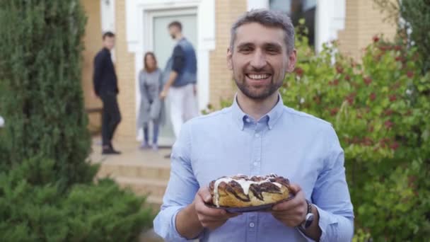 Lächelnder Kaukasier, der mit einer Torte steht und lächelt. eine Gruppe von Menschen im Hintergrund, die miteinander reden. Nachbarn verbringen Zeit gemeinsam im Freien. — Stockvideo