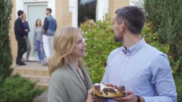 Ευτυχισμένο ώριμο ζευγάρι με κέικ χαμογελά και κοιτάζοντας την κάμερα στέκεται στο προσκήνιο, ενώ η παρέα των ανθρώπων που μιλούν στη βεράντα στο παρασκήνιο. Γνωρίστε νέους γείτονες — Αρχείο Βίντεο