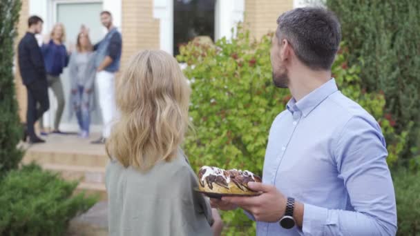 Ευχάριστο ώριμο ζευγάρι με κέικ χαμογελά και κοιτάζοντας πίσω σε μια ομάδα των ανθρώπων που χαιρετίζουν. Καυκάσιος οικογένεια χαιρετά νέους γείτονες με ένα δώρο. — Αρχείο Βίντεο