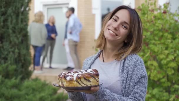 Junge attraktive Frau mit einer Torte, die lächelt und in die Kamera blickt. Gesellschaft von Menschen, die sich auf der Veranda im Hintergrund unterhalten. Neue Nachbarn kennenlernen. — Stockvideo