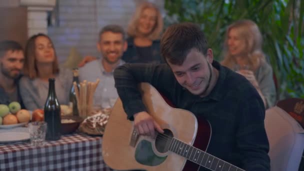 Compañía de personas bebiendo alcohol y sonriendo sentados a la mesa en el fondo. Joven tocando la guitarra en primer plano. Amistosa familia celebrando vacaciones — Vídeos de Stock