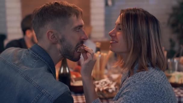 Gesellschaft fröhlich lächelnder Menschen, die im Hintergrund am Tisch sitzen, während im Vordergrund eine junge Frau ihren Mann mit einem Sandwich füttert. freundliche Familie feiert Feiertag — Stockvideo