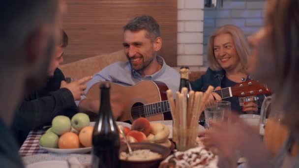 Зрелый кавказский человек играет на гитаре в компании друзей. Счастливая компания проводит время вместе вечером. Соседи поют и празднуют праздник . — стоковое видео