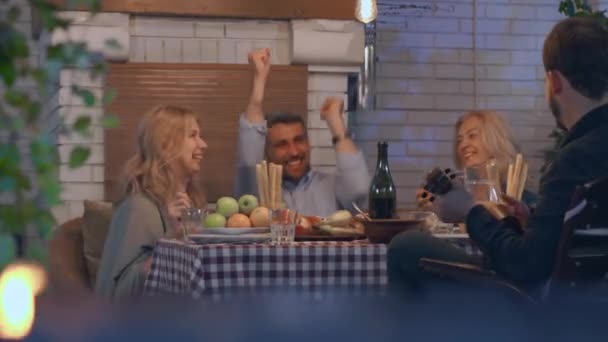 Compañía de gente bebiendo alcohol y sonriendo sentados a la mesa. Un joven tocando el ukelele, sus amigos bailando. Amistosa familia celebrando vacaciones — Vídeos de Stock