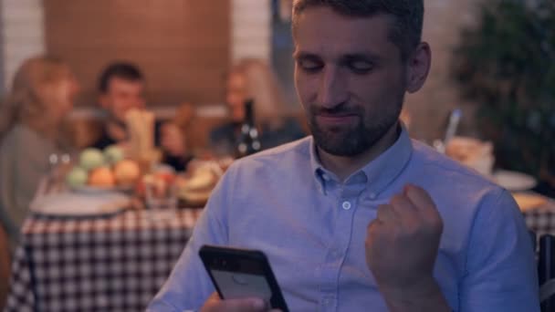 Volwassen blanke man zittend op de voorgrond spelen op de telefoon met zijn terug naar het gezelschap van jongeren vieren vakantie. Verslaving aan games, apparaten en sociale media. — Stockvideo