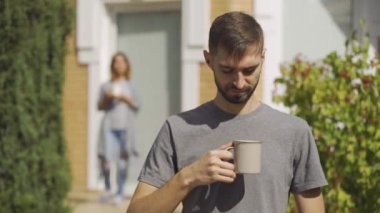 Yakışıklı adam ön planda çay bardağı ile duran kameraya bakarak, kız arka planda duran. Evde genç bir aile var. Yaz.