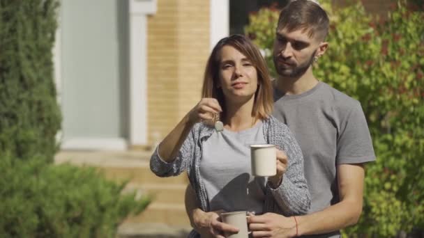 Retrato alegría mujer caucásica con la taza de té y hombre guapo de pie delante de la casa grande. Llave de la casa de exposición. Propiedad . — Vídeos de Stock