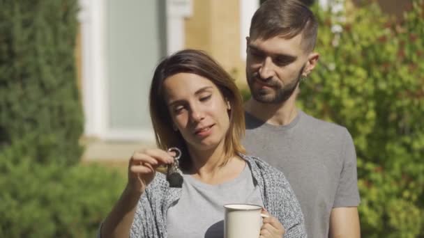 Портрет щасливої білої жінки з чашкою чаю і красивим чоловіком, що стоїть перед великим будинком. Дівчина показує ключ будинку. Власність . — стокове відео