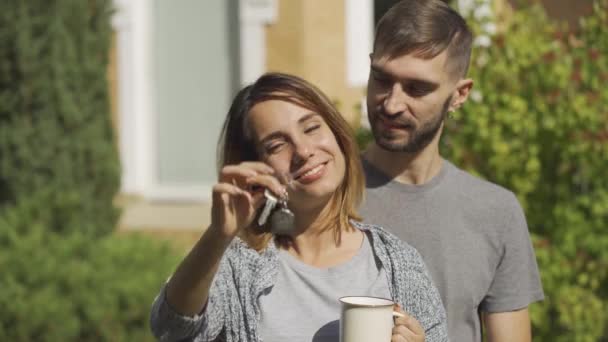Portræt af attraktiv hvid kvinde med kop te og flot mand stående foran et stort hus. Konen viser husnøgle og smiler. Ejerskab – Stock-video
