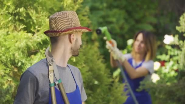 Красивий молодий фермер у блакитному халаті стоїть на передньому плані, а його дружина поливає дерева на задньому плані. Чоловік і жінка працівник в саду — стокове відео