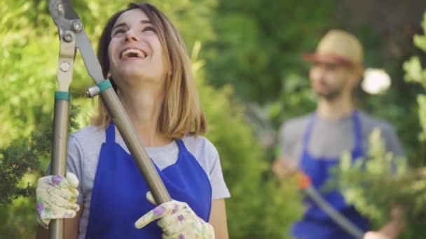 Retrato de una linda mujer caucásica mostrando el pulgar hacia arriba sosteniendo un gran cortador de jardín en primer plano mientras el hombre riega árboles en el fondo. Trabajador y trabajadora en el jardín — Vídeos de Stock