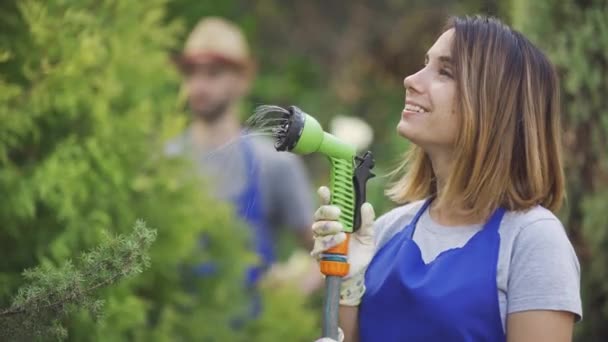 Retrato de una hermosa mujer caucásica jugando con una manguera de agua en primer plano mientras el hombre corta hojas de árbol en el fondo. Trabajador y trabajadora en el jardín — Vídeo de stock