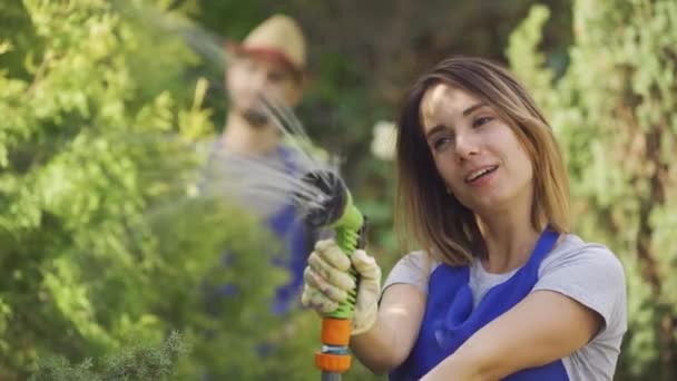 Portrét přitažlivé kavkazské ženy, která si hraje s vodní hadicí v popředí, zatímco člověk střímá listí na pozadí. Mužský a ženský pracovník v zahradě — Stock video