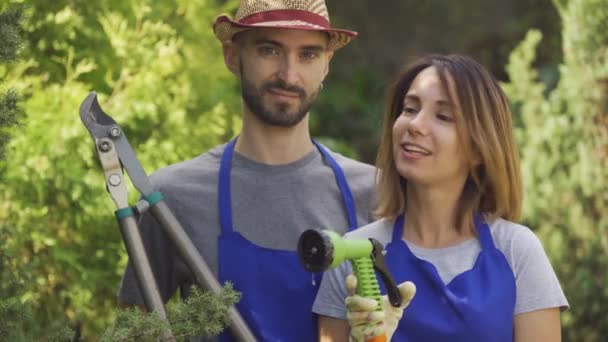 Retrato sonriente mujer caucásica y hombre guapo en uniforme azul de pie en el jardín sosteniendo herramientas de jardín mirando a la cámara. Trabajadores y trabajadoras en el jardín. Familia joven de agricultores — Vídeo de stock