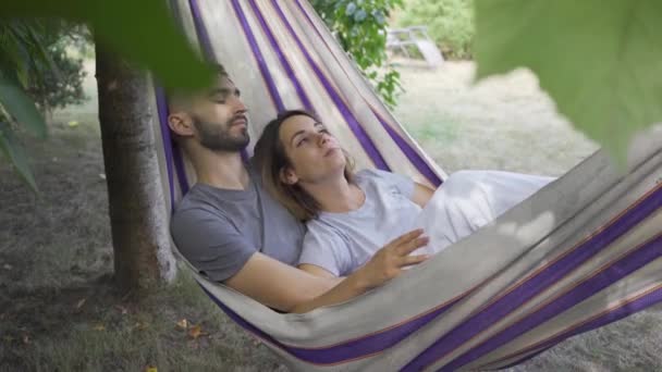 Junge kaukasische Mann und Frau liegen in der Hängematte im Garten und entspannen zusammen. Liebespaar zusammen im Freien. Sommerfreizeit — Stockvideo
