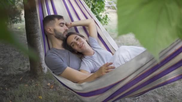 Cute young caucasian man and woman lying in hammock in the garden relaxing together. Loving couple together outdoors. Summertime leisure — Stock Video