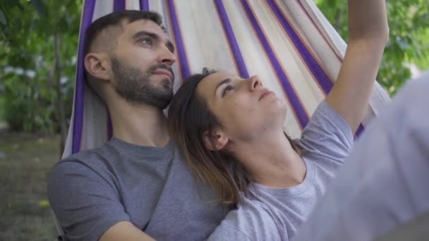 Feliz joven caucásico hombre y mujer acostados en hamaca en el jardín relajándose juntos. Amar a la pareja al aire libre. Tiempo libre de verano — Vídeos de Stock