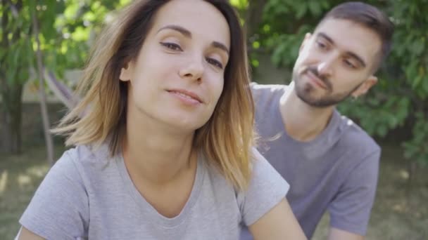 Portrait of young man and woman relaxing in summer garden. The guy touching hair of his wife, she smiling looking at camera. Happy young family resting outdoors. Concept of relationship, tenderness — Stock Video