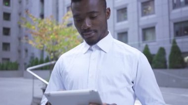 Şehir caddesinde duran tablet ile çalışan beyaz gömlekyakışıklı Afro-Amerikan adam portresi. Modern binada erkek işadamı veya yöneticisi