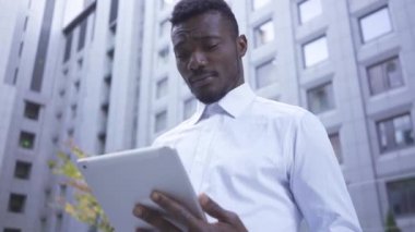 Endişeli erkek Afro-Amerikan beyaz gömlek li tablet gökdelenlerin arka planda duran ile çalışıyor. Modern binada erkek işadamı veya yöneticisi.