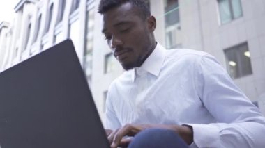 Kendine güvenen Afro-Amerikan adam beyaz gömlekli dizüstü bilgisayar da şehir caddesinde oturan yazarak. Dışarıda çalışan erkek işadamı veya yönetici.