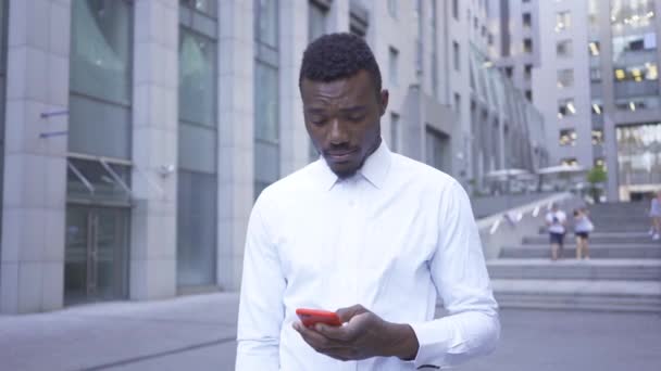 Стурбований афро-американської людини в білій сорочці говорити червоний стільниковий телефон стоячи на міській вулиці. Чоловічий бізнесмен або менеджер поруч з сучасним будівлею. — стокове відео