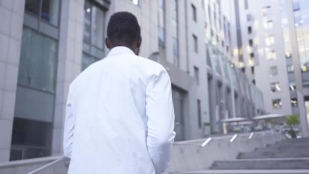 Vista posterior del hombre afroamericano en camisa blanca caminando hacia un gran centro de negocios moderno. Hombre de negocios o gerente que va a la oficina — Vídeos de Stock