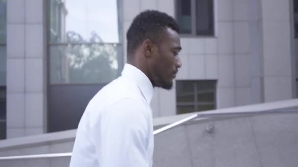Zijaanzicht van Afro-Amerikaanse man in wit shirt wandelen op de trap in de buurt van het grote moderne businesscentrum. Mannelijke zakenman of manager naar kantoor. — Stockvideo