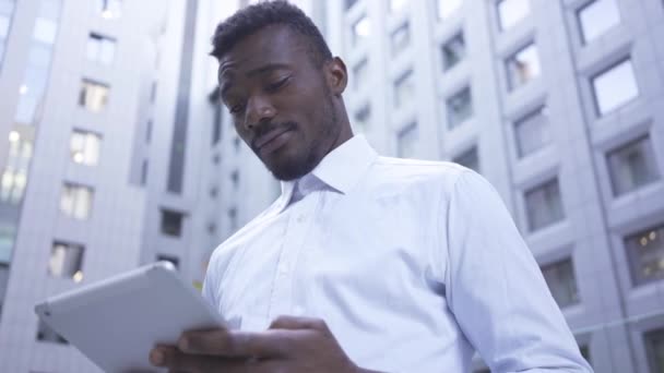 穿着白衬衫的深思熟虑的非洲裔美国人，站在摩天大楼的背景上，与平板电脑一起工作。现代建筑的男性商人或经理. — 图库视频影像