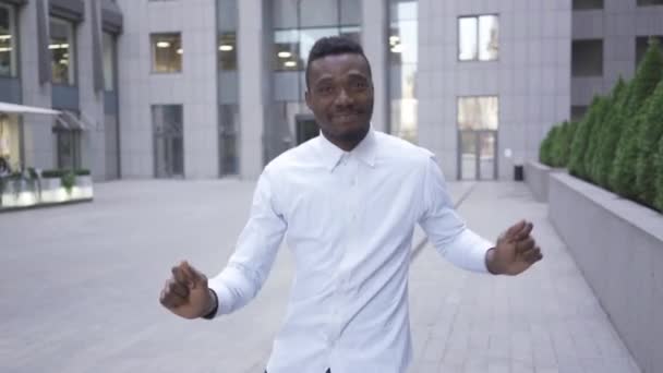 Счастливый афроамериканец в белой рубашке танцует перед современным бизнес-центром на городской улице. Мужчина бизнесмен или менеджер получил отличные новости — стоковое видео