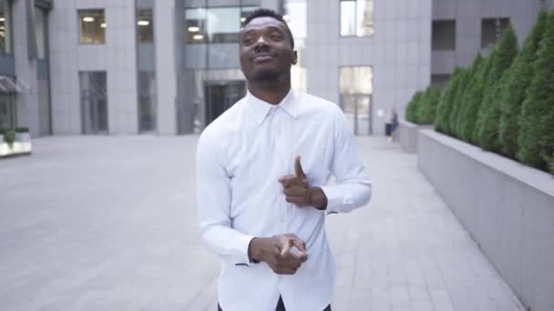 Hombre afroamericano satisfecho con camisa blanca bailando frente al moderno centro de negocios en la calle de la ciudad. Hombre de negocios o gerente consiguió grandes noticias . — Vídeos de Stock