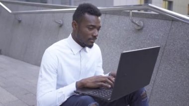 Beyaz gömlekli Pozitif Afro-Amerikan adam şehir caddesinde oturan dizüstü bilgisayarda yazıyor. Dışarıda çalışan kendinden emin erkek işadamı veya yönetici.