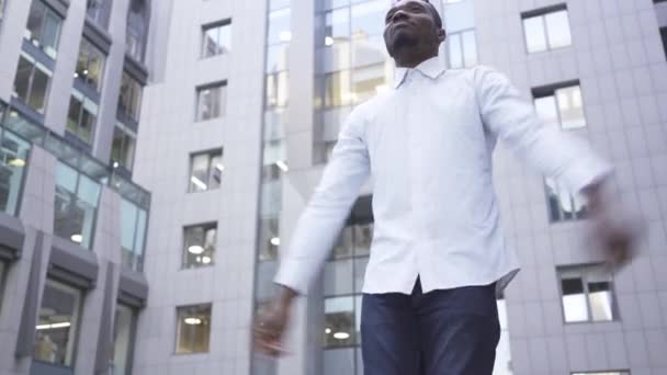 Нижний вид счастливого афроамериканца в белой рубашке, танцующего перед современным бизнес-центром на городской улице. Мужчина бизнесмен или менеджер получил отличные новости . — стоковое видео