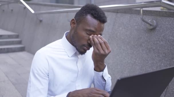 Hombre afroamericano exhausto con camisa blanca frotándose los ojos y escribiendo en el portátil. Hombre cansado hombre de negocios o gerente trabajando al aire libre . — Vídeos de Stock