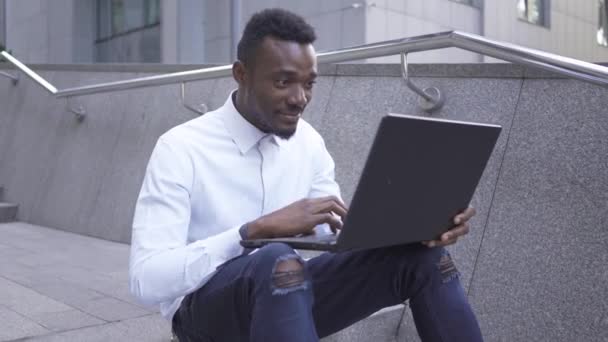 Портрет красивого афроамериканца в белой рубашке, печатающего на ноутбуке, сидящего на городской улице. Парень делает победный жест. Уверенный мужчина-бизнесмен или менеджер, работающий на открытом воздухе — стоковое видео
